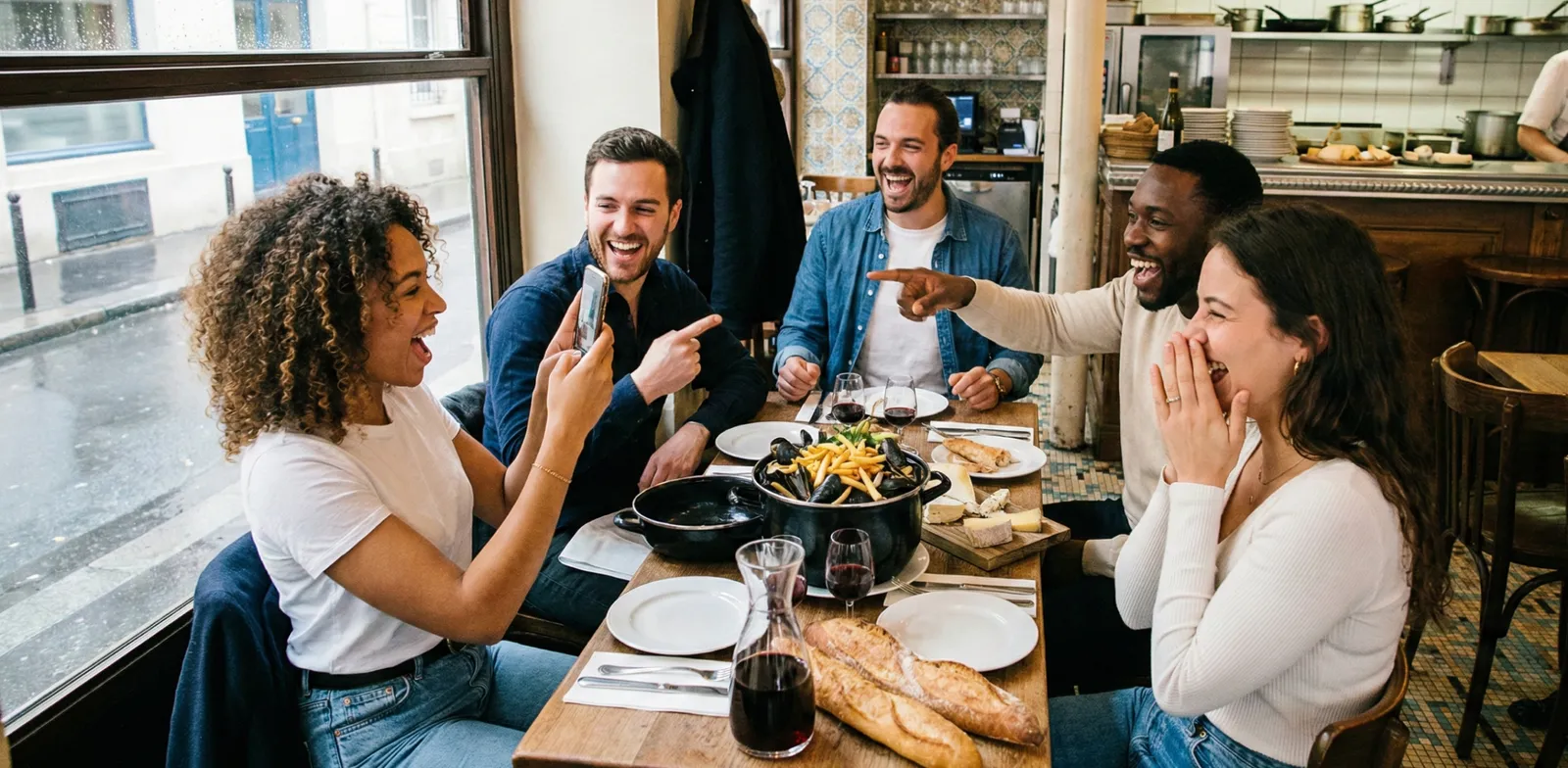Cinco personas ríen en la mesa de un restaurante mientras una de ellas levanta su celular.